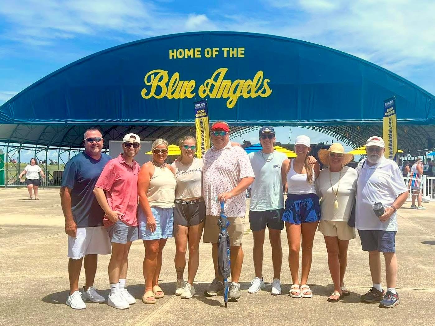 A Group of Seven People Stands in Front of a Large Blue Canopy Labeled “Home of the Blue Angels,” Enjoying a Sunny Day at an Airshow Event.