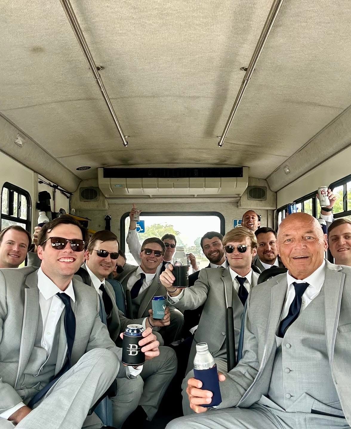 Groom and Groomsmen Celebrate on a Bus, All Dressed in Matching Gray Suits, Holding Drinks and Smiling Joyfully. The Scene Captures Pre-Wedding Festivities.