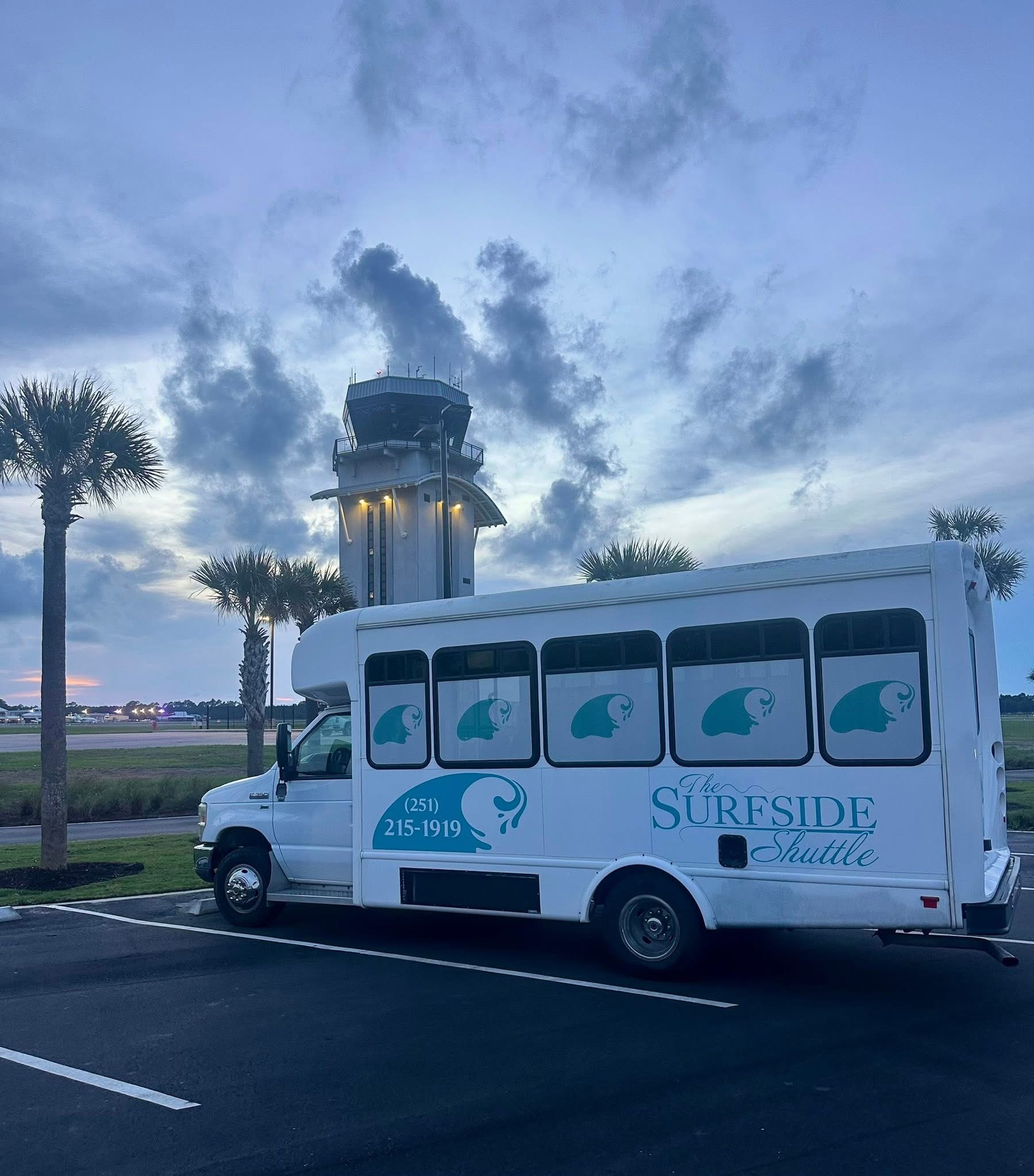 Sureside Shuttle Bus Parked Near an Airport Tower at Dusk, With Palm Trees and Cloud Patterns in the Background, Emphasizing Transportation Services.