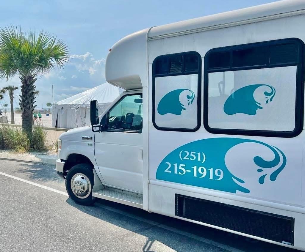 A Shuttle Bus Parked Along a Palm-Lined Road Features Blue Wave Graphics and a Contact Number. It Suggests Transportation Services Near the Beach.